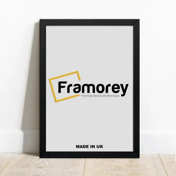 Ash Black picture frame with a Framorey Logo place inside the Frame and the frame is Standing on a wooden surface, leaning with the wall.